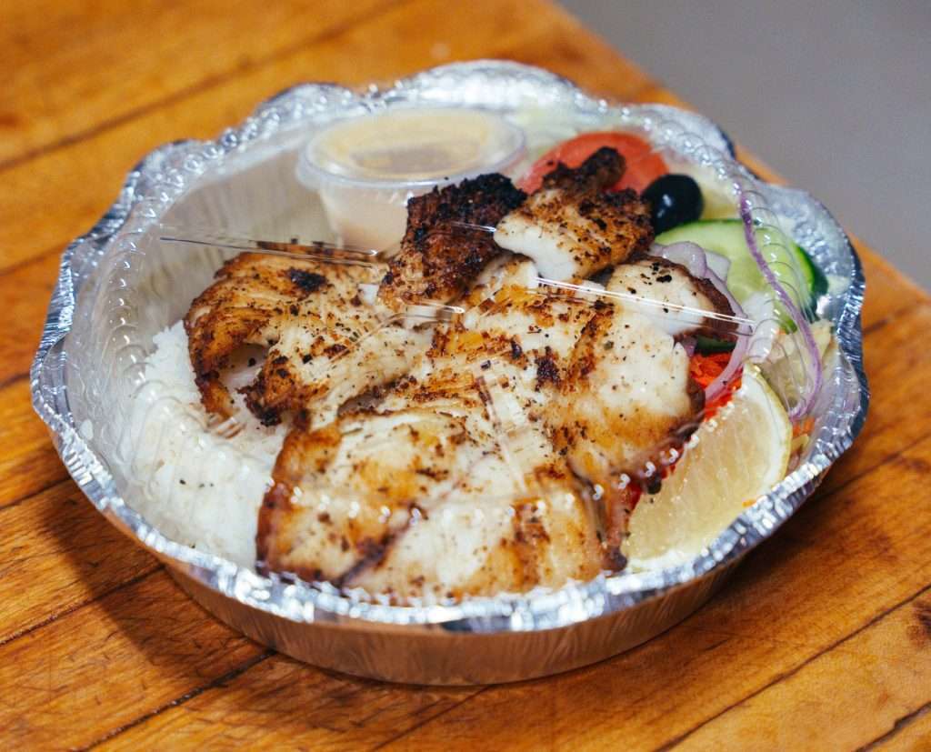 A to go order of grilled chicken over white rice, with vegetables and a slice of lemon.
